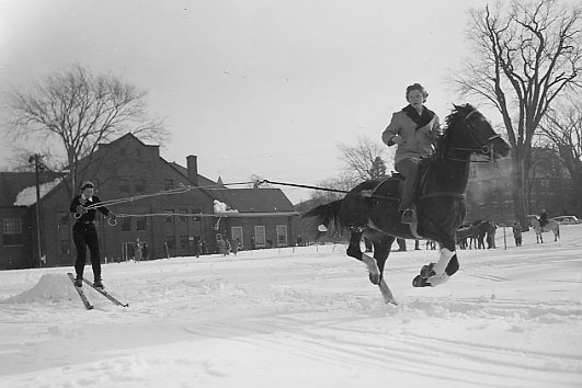 A women riding a horse in the snow pulls another figure on skis