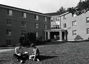 Entrance to Lord Hall with woman exiting and two people sitting on lawn in foreground.