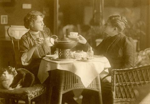 Amy Beach having tea with her friend and a cat sits nearby